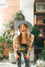 Wearing the Astral Connection Festie Tank, a woman with long wavy hair and a large hat smiles brightly, leaning forward in patterned pants amidst a room filled with plants, books, and a vintage TV.