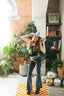 A woman smiles as she poses in a stylish room filled with plants, disco balls, a retro TV, and a bookshelf on a checkered rug. Sunlight streams through the window as she wears a blue hat, patterned pants, and the Astral Connection Festie Tank.