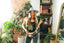 A smiling woman wearing the Astral Connection Festie Tank, patterned pants, and a hat poses indoors making a peace sign. She stands among green plants, bookshelves, and a mirror with sunlight streaming in.