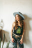 A smiling woman in the Astral Connection Festie Tank, a wide-brimmed hat, and patterned pants stands by a wall with plants nearby.