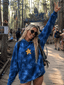 Wearing the Stargaze Tie Dye Hoodie, a smiling woman flashes a peace sign at an outdoor festival in a forest, with people and a Shambhala sign visible in the background.