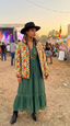 A woman wearing a floral print jacket, green dress, and black hat stands in a field with a festival atmosphere in the background.