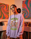 A person with long braids and hoop earrings wears the Bad Bitches Love Bass Crewneck, a lavender vintage-style sweatshirt featuring a colorful butterfly and bold “Bad Bitches Love Bass” text. They stand in front of psychedelic vinyl records and vibrant album art, making this crewneck the perfect statement piece for festival clothing.