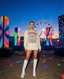 Wearing the Disco Tribe Festie Club Crewneck and white boots, a woman stands outdoors at night among colorful LED pillars, with a glowing Ferris wheel in the background, creating a vibrant festival vibe.