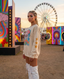Wearing the Disco Tribe Festie Club Crewneck and white boots, a woman stands at a festival surrounded by vibrant art installations and a Ferris wheel. With braided hair and glittery face paint, she gazes toward the camera at sunset.