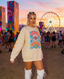 A woman with braided hair and vibrant face paint wears the Disco Tribe Festie Club Crewneck paired with white boots at a lively outdoor festival, featuring a ferris wheel and art installations in the background during sunset.