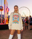A woman with blonde braids, wearing the Disco Tribe Festie Club Crewneck and white boots, stands at a festival with a Ferris wheel and vibrant lights illuminating the sunset sky in the background.