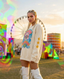 Wearing the Disco Tribe Festie Club Crewneck, a woman with braids and face glitter stands outdoors at a festival, framed by colorful art installations, a Ferris wheel, and a vibrant sunset sky.