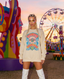 A woman with colorful face gems and braids stands at a festival, wearing the Disco Tribe Festie Club Crewneck and white boots. Bright sculptures and a Ferris wheel are in the background at sunset.