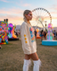 Wearing the Disco Tribe Festie Club Crewneck and white boots, a woman with braided hair and glitter makeup stands at a festival. Behind her, vibrant art installations and a large Ferris wheel glow beneath the sunset sky.