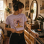 A young woman wearing a purple crop top with a mushroom graphic and the text "Disco Tribe" is standing in a room with a record player and various decor items.
