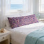 a serene bedroom scene with a large bed adorned with a purple and pink patterned pillow, a white comforter, and a blue blanket. The bed is positioned next to a window that offers a view of the ocean, creating a tranquil and relaxing atmosphere.