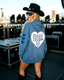 A woman wearing a blue hoodie with a heart design and a black hat stands in front of a stage with various equipment and speakers.